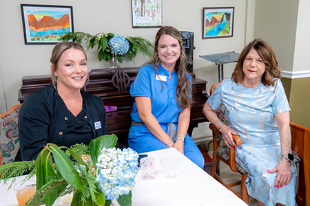 Three Diversicare healthcare team members smiling together at a table, wearing medical scrubs and a light blue dress.