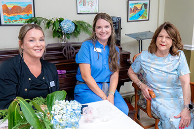 Three Diversicare healthcare team members smiling together at a table, wearing medical scrubs and a light blue dress.